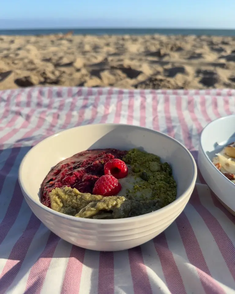Pistachio Addiction Bowl with raspberry-chia pudding, whole raspberries, crushed pistachios and pistachio butter on a pink striped beach towel